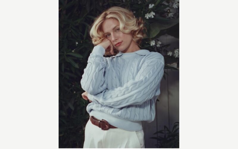 Ashe wearing a pale sweater in front of green foliage.
