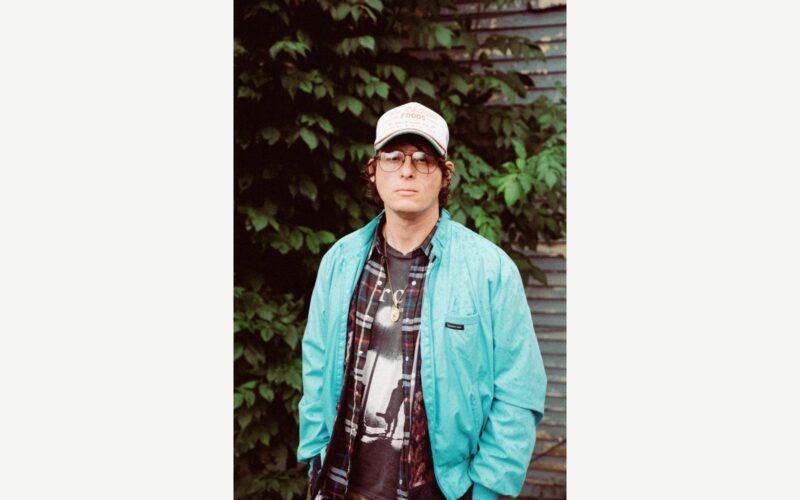 Stephen Wilson Jr. wearing a baseball cap, glasses and a jacket in front of green foliage.
